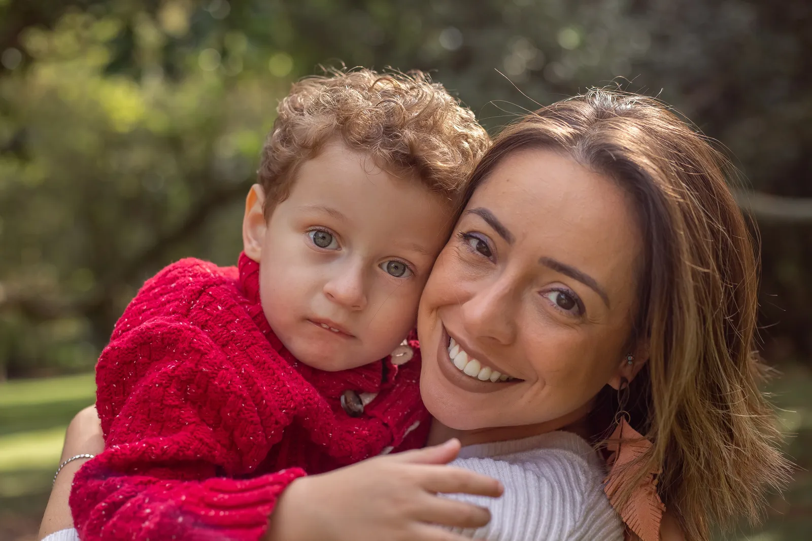 A smiling woman holds a curly-haired little boy cheek to cheek in a park.